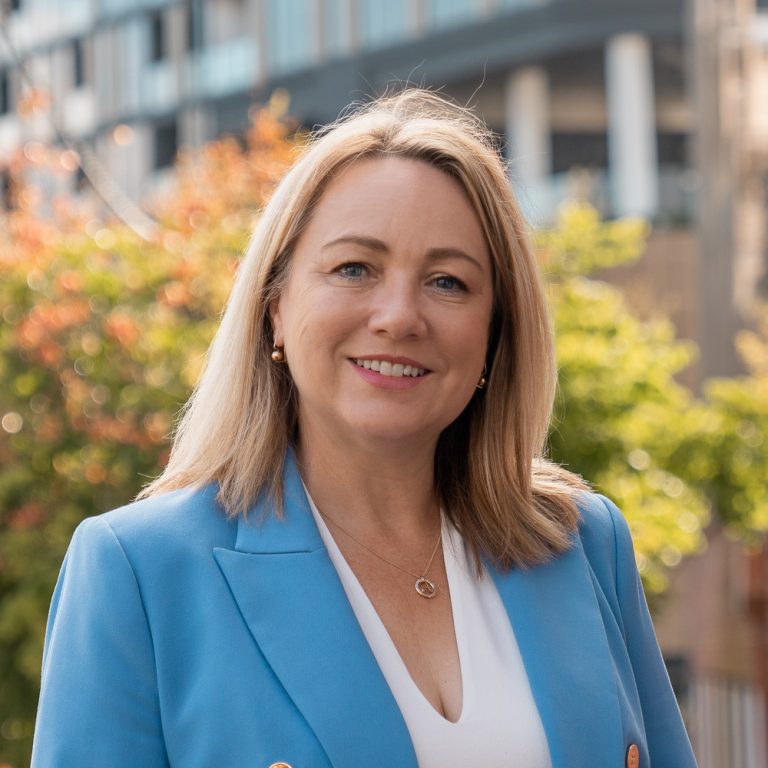 Alisa Cork. A middle aged woman with dyed blond hair. Wearing a white blouse and bright blue suit jacket.