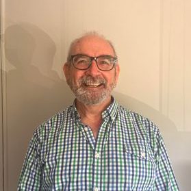 Jim Haywood, an older white man with a white beard, large glasses and a checkered button-up shirt. He is grinning at the camera with a warm, welcoming smile.
