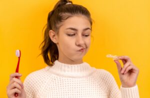 A teenage girl holding a toothbrush in one hand a teeth guard in the other hand. She is pulling a face while looking at the teeth guard.