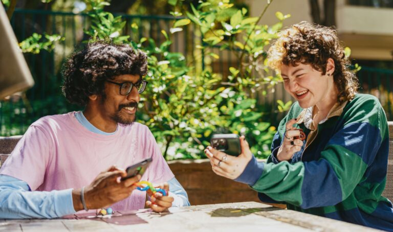 A young Autistic man sits at an outdoor table with a young Autistic woman who is showing him her mobile phone. They are both smiling as they build social connections and independence in the community.