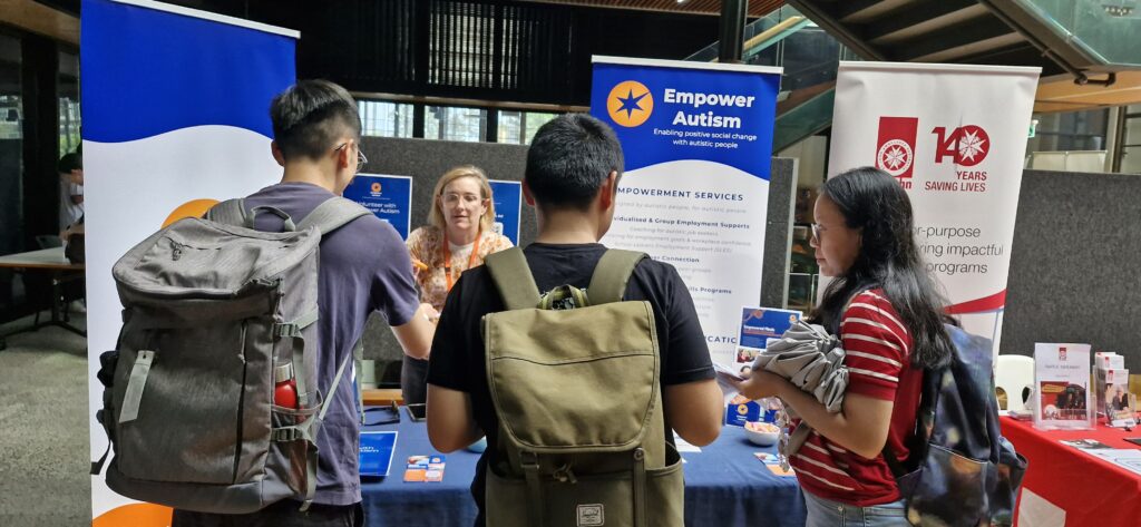 Empower Autism branded table at a community convention. A middle aged caucasian woman stands behind the desk, sharing information with three young adult visitors.
