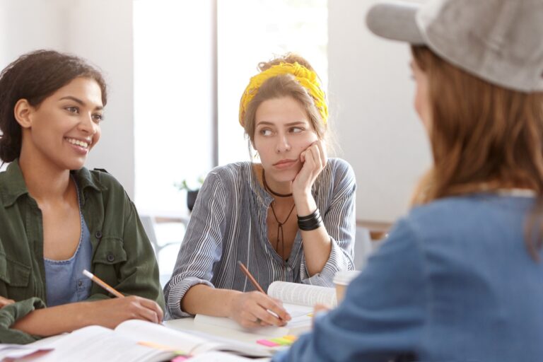 A work meeting between three young women. Two of the women smile and collaborate with one another, while the third watches silently.