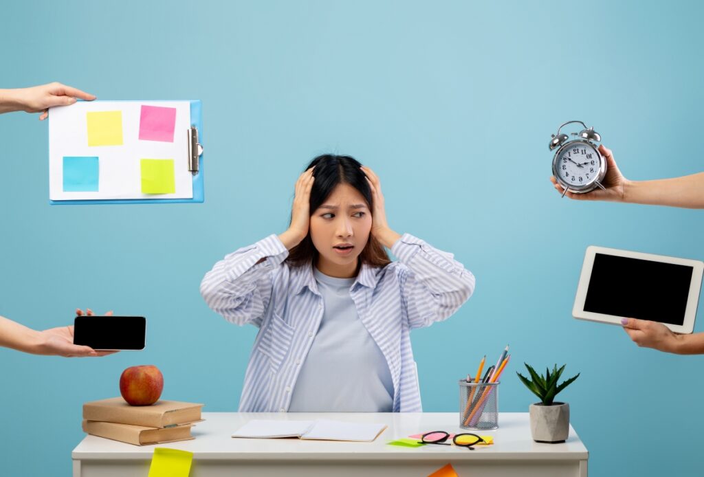 A young South-East Asian woman sits at an office desk. From outside of the photo's frame, hands reach in to hold various items of responsibility towards her; a clipboard, an alarm clock and a phone. She looks at the items with hands over her ears, distressed.