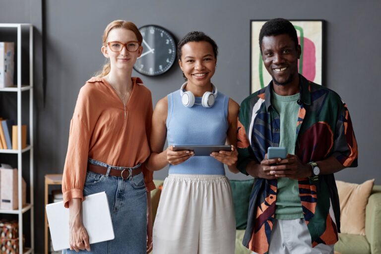 Three young people in colourful, semi-casual workwear stand together in a collaborative work space, smiling.