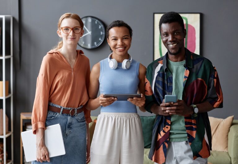 Three young people in colourful, semi-casual workwear stand together in a collaborative work space, smiling.
