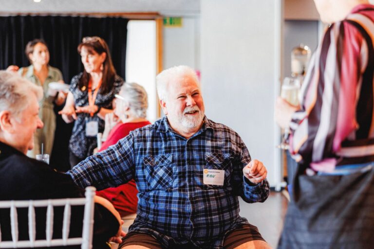 An older man with white hair and beard, talking to a small group of other Autistic adults. He has a small, Empower Autism branded, name tag which reads "Hello, my name is Ron."