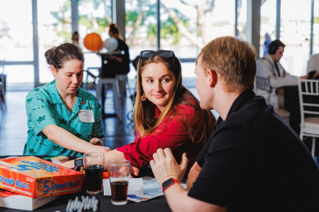 Three young adults sit at a table covered in boardgames. One man turns to speak to a younger woman who looks at him, smiling.