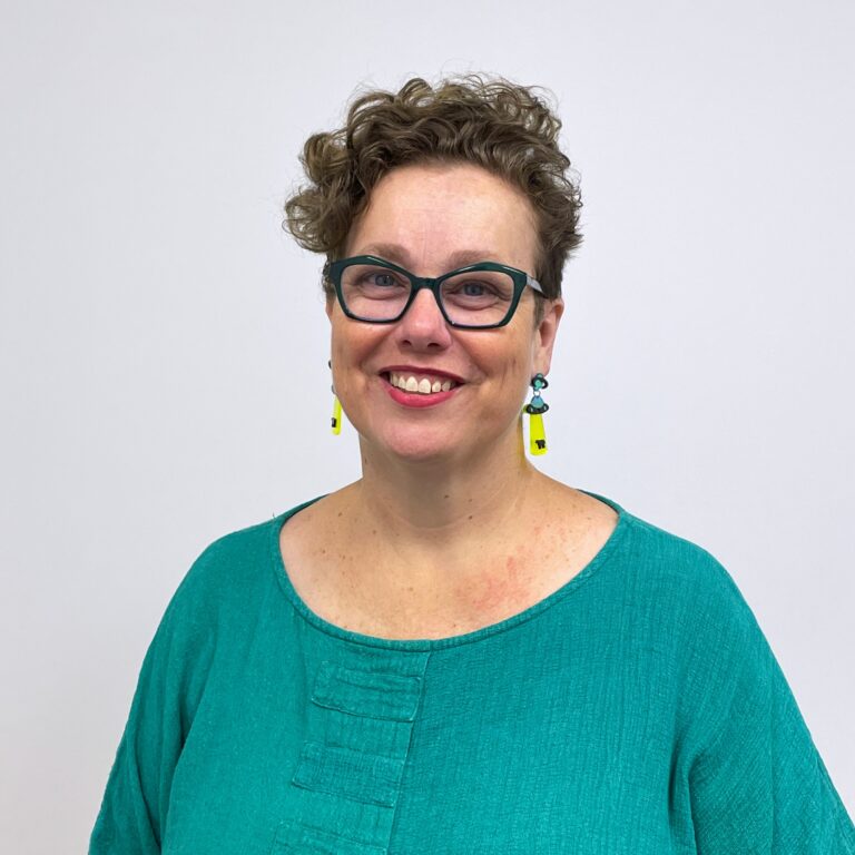 Nerine Broome. A middle aged woman with short curly brown hair, wearing a green linen blouse, chunky green glasses and green and yellow dangly UFO earrings.