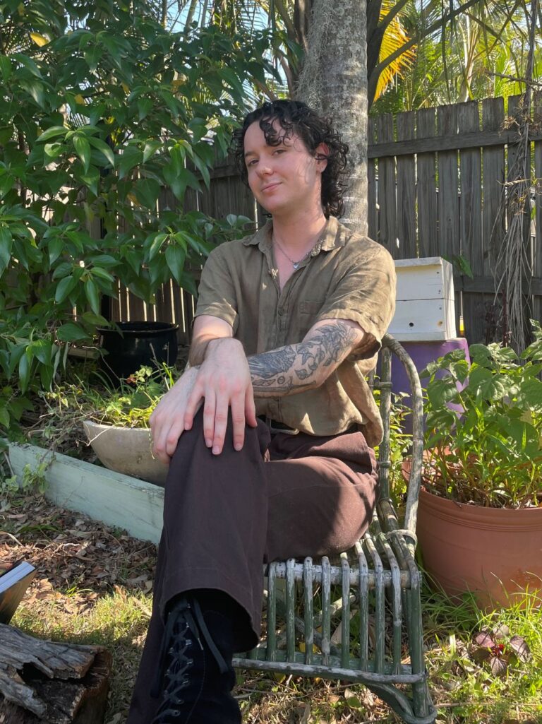 Riley, a young white man with curly hair and a patchwork tattoo sleeve, sitting on a wicker chair in a lush home garden.