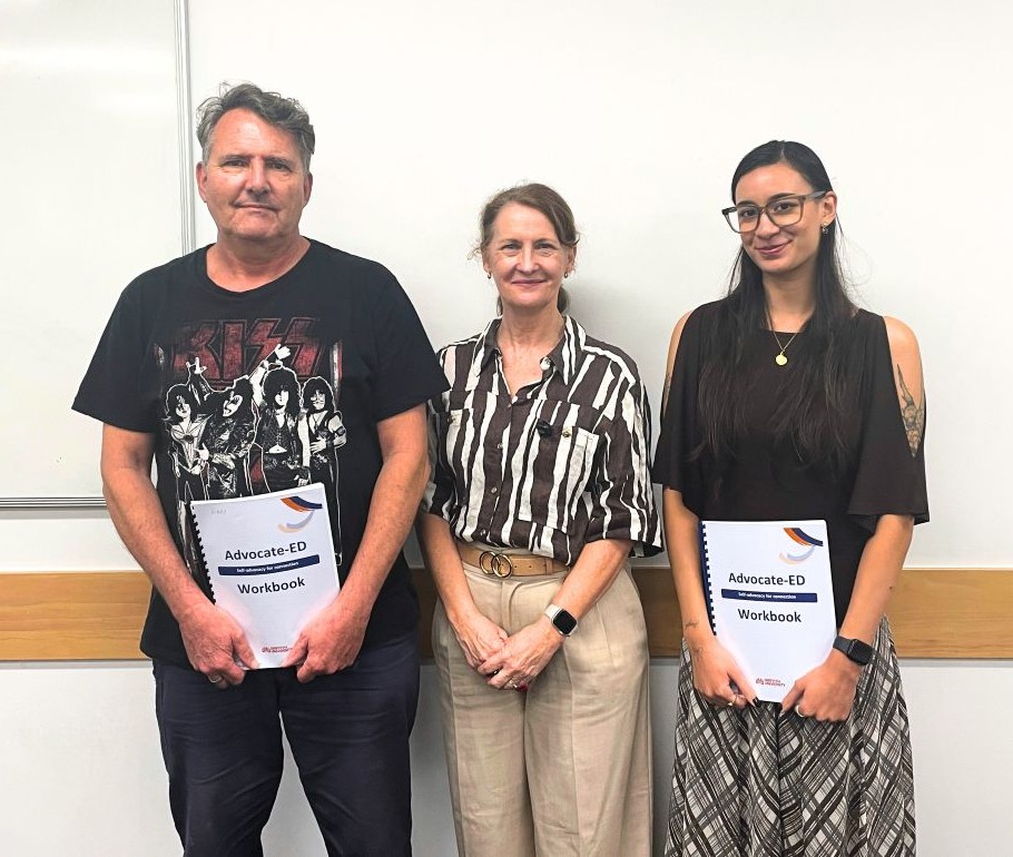 Two univsity students, an older white man and a young south-east asian woman, stand beside the Advocate-ED facilitator, holding Advocate-ED booklets.
