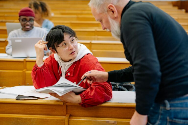 A university tutor, an older white man with a beard, assists an east-asian student in his early 20s with his study.