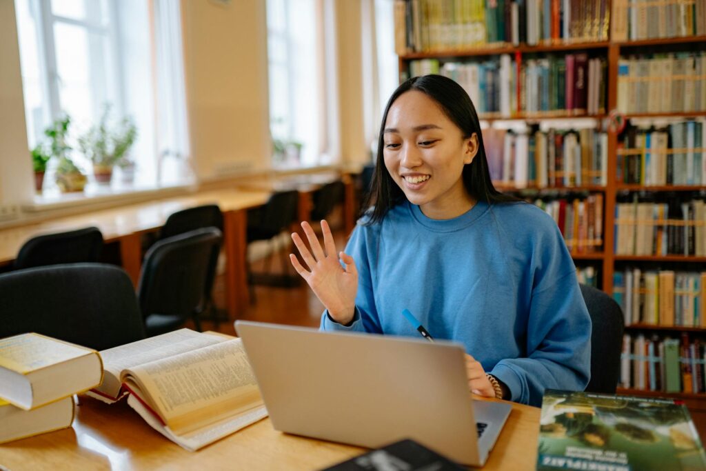 A tertiary student, a young south-east asian woman, greets her online class, sitting in a library.