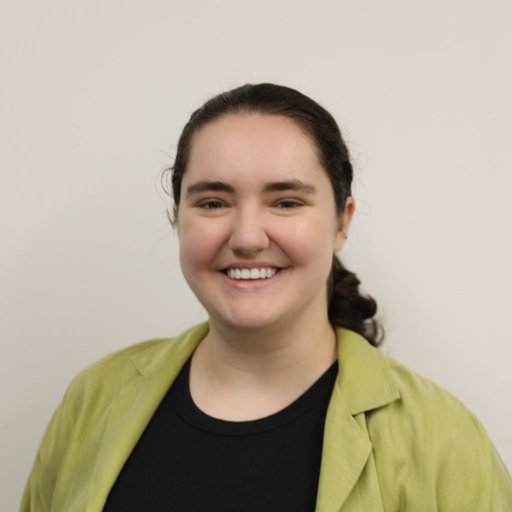 Photograph of Elise Manson, a young woman with a slicked back pony tail and a bright green jacket, grinning.