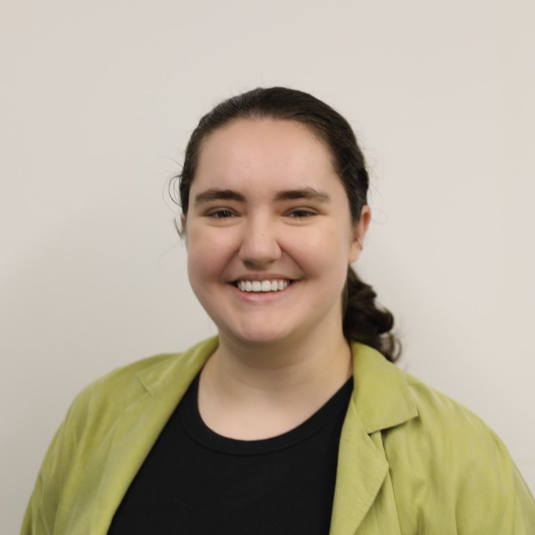 Photograph of Elise Manson, a young woman with a slicked back pony tail and a bright green jacket, grinning.