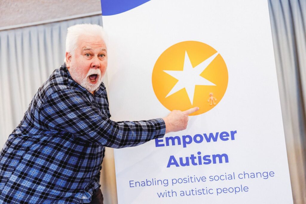 A senior white man with short white hair and beard points to an Empower Autism banner enthusiastically.