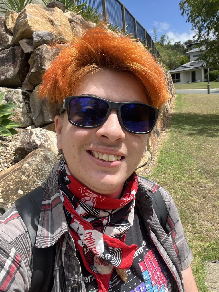 Selfie of Forrest, a young trans man with dyed red hair; wearing sunglasses and a playful patterned shirt, at a park.