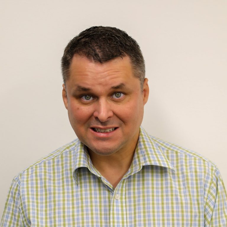 Photograph of Michael, a middle aged white man, smiling and wearing a plaid button-up shirt.