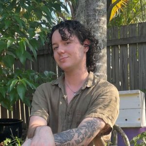 Photograph of Riley Sellick, a young white trans man white curly dark hair and a patch-work tattoo sleeve, sitting in a leafy garden.