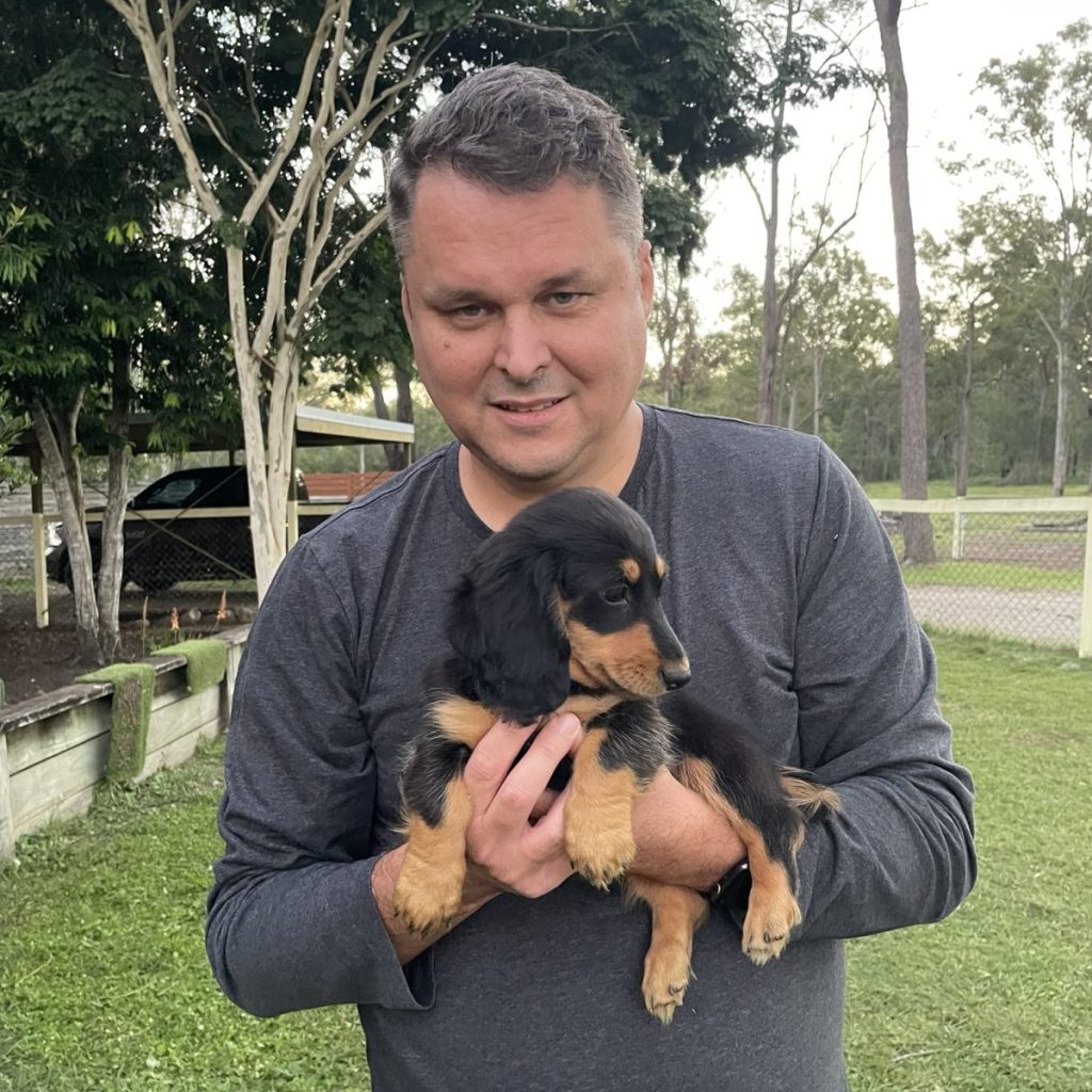 Michael standing in a park, gently holding a long haired dauschound dog and smiling.
