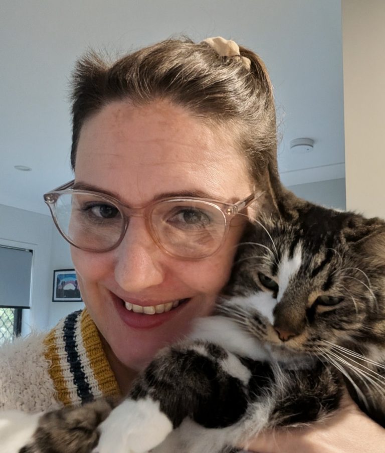 Selfie of Jac Lai, a white woman with short hair and large-framed glasses, hugging a very relaxed, longhaired tabby cat.