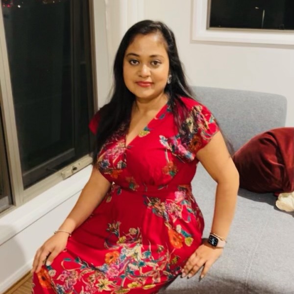 Rani Sinha, an Indian woman in her thirties with long straight hair and bright red lipstick, sitting on the edge of a couch, wearing a long, flowy red floral dress.